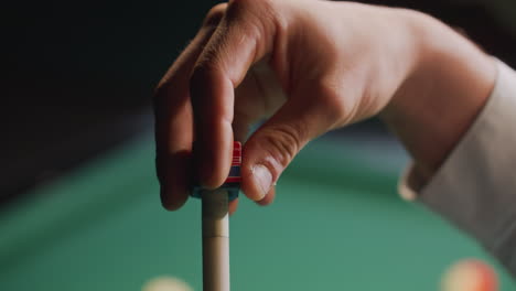 close-up of player's hand applying chalk to cue stick before executing precise shot on green pool table. focus on preparation and technique with blurred billiard balls in background