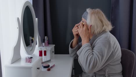 Old-senior-woman-grandmother-taking-care-of-skin-near-eyes-and-wrinkles,-putting-makeup-on-at-home