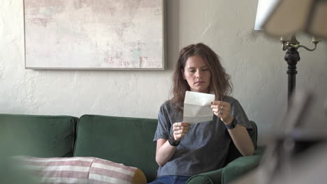 a woman sits on a green couch, deeply focused on a love letter. the camera moves sideways, shooting behind a lamp and plants, creating a beautiful parallax effect.