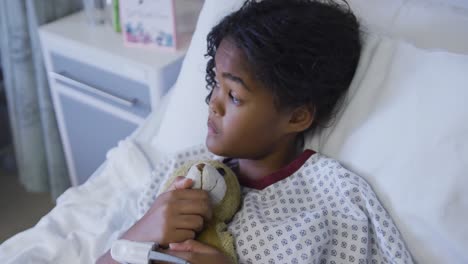 Mixed-race-girl-lying-on-hospital-bed-wearing-fingertip-pulse-oximeter-and-holding-teddy-bear