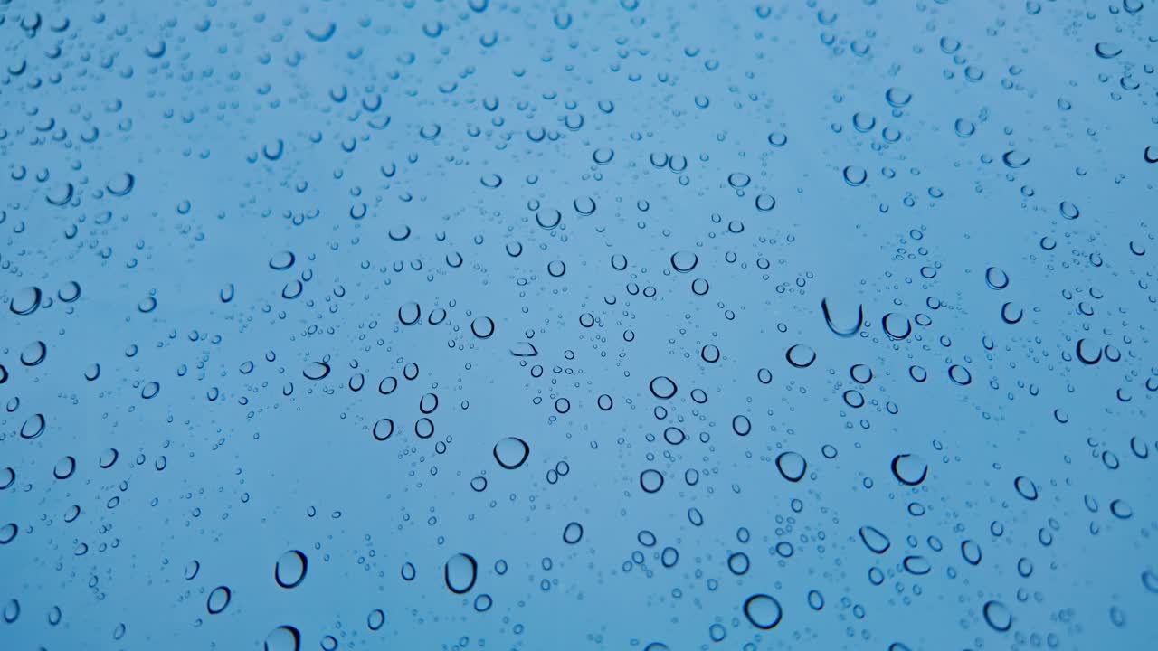 Nahaufnahme von Regentropfen auf Glasoberfläche mit kühlen Blautönen, bewölktes Regenwetter.