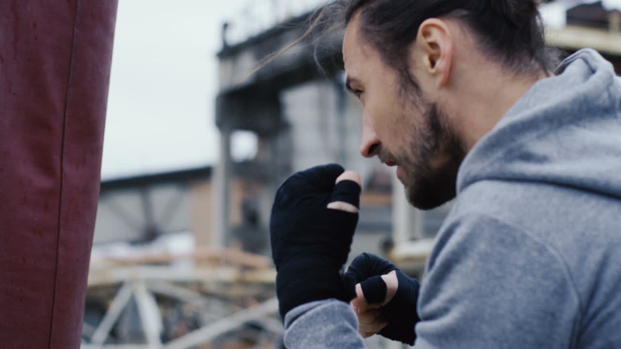 vista de cerca de un hombre caucásico con ropa deportiva golpeando un saco de boxeo al aire libre en una fábrica abandonada en una mañana nublada