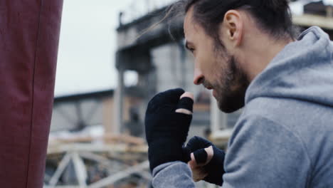 vista de cerca de un hombre caucásico con ropa deportiva golpeando un saco de boxeo al aire libre en una fábrica abandonada en una mañana nublada