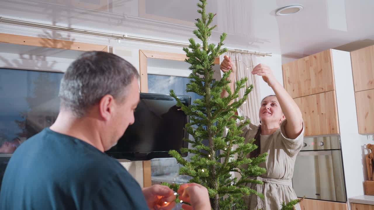 Woman in brown gown fixing Christmas light on festive tree while husband in green top assists her in decorating for the holiday season, working together in a joyful festive atmosphere