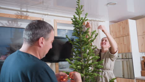 woman in brown gown fixing christmas light on festive tree while husband in green top assists her in decorating for the holiday season, working together in a joyful festive atmosphere