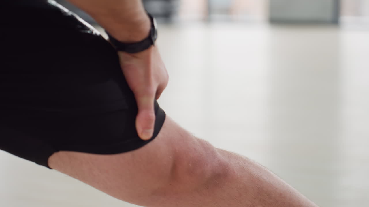 Trainer hands on thigh adjusting position during deep side lunge stretch in modern bright gym room wearing white tank top and black shorts on light wooden floor with large windows backdrop