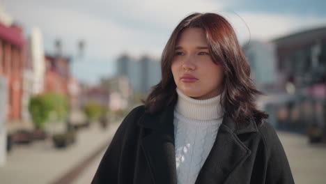 confident woman in black coat walking outdoors, looking thoughtful looks around, with blurred background showing buildings, people walking, and a city statue