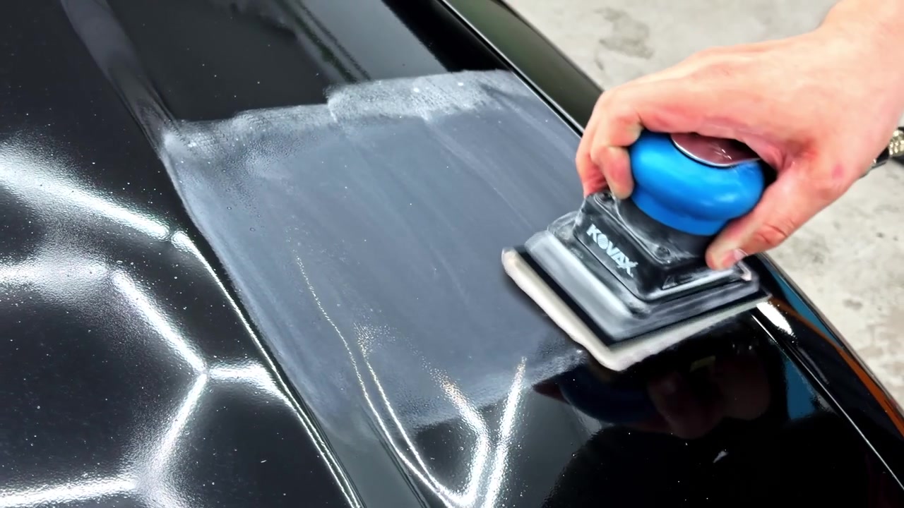 Close-up of a hand using a pneumatic sanding block on a black car panel, showing paint correction, body preparation, and professional auto detailing work.