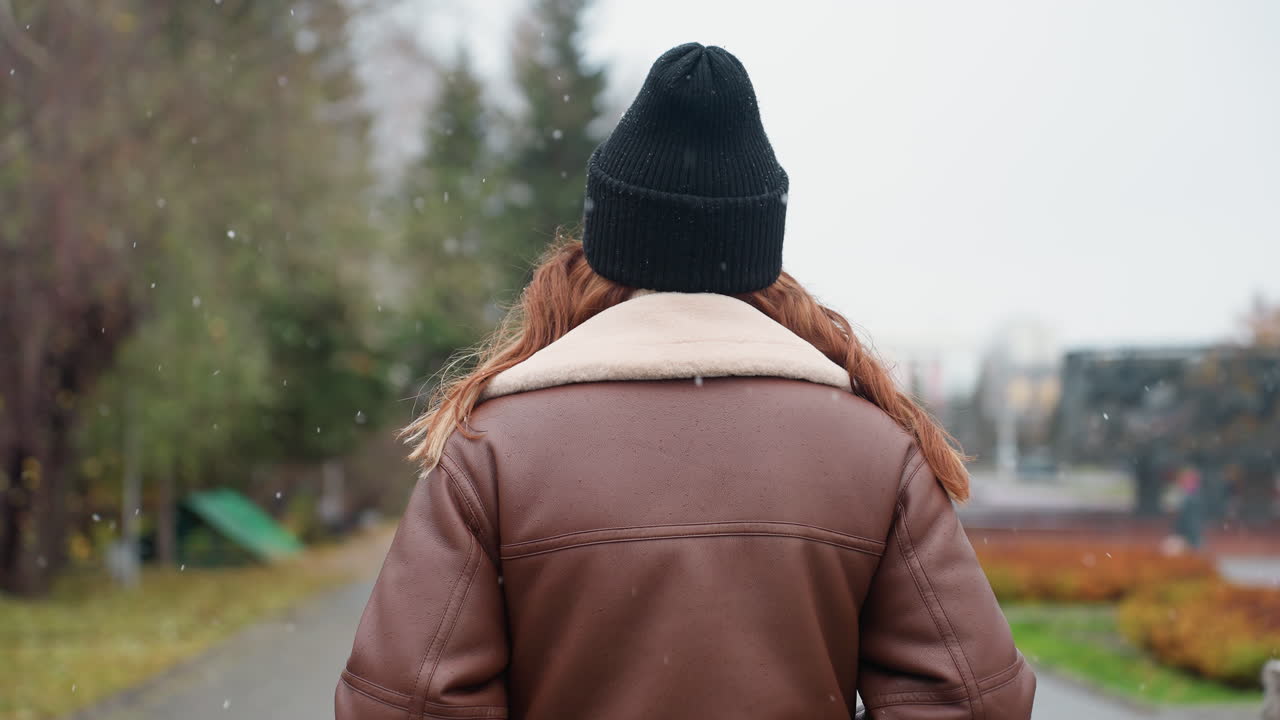 Back view of young woman in black cap and brown shearling jacket walking slowly with hands in pockets on wide stone pathway through city park during calm snowy day