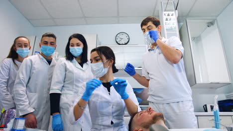 female brunette student takes the metal wire out off the patient's mouth. practicing student takes the tool from stand. group of colleagues at backdrop. low angle view.