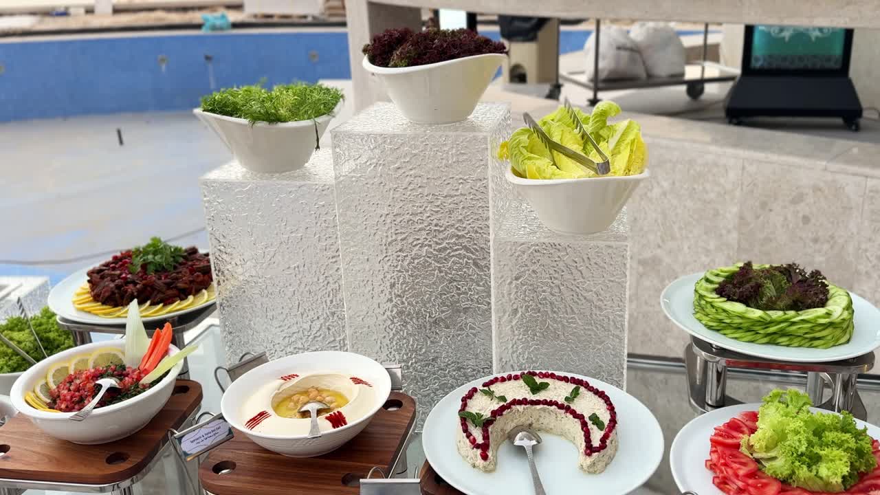 Elegante salad bar all'aperto con hummus, insalate miste, pomodori a fette, cetrioli, tabulé e vassoi di verdure artisticamente disposti su supporti moderni vicino alla piscina.