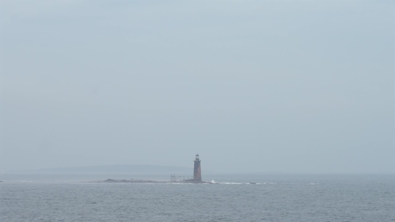 A small lighthouse in the harbor with waves crashing around it on a foggy day.