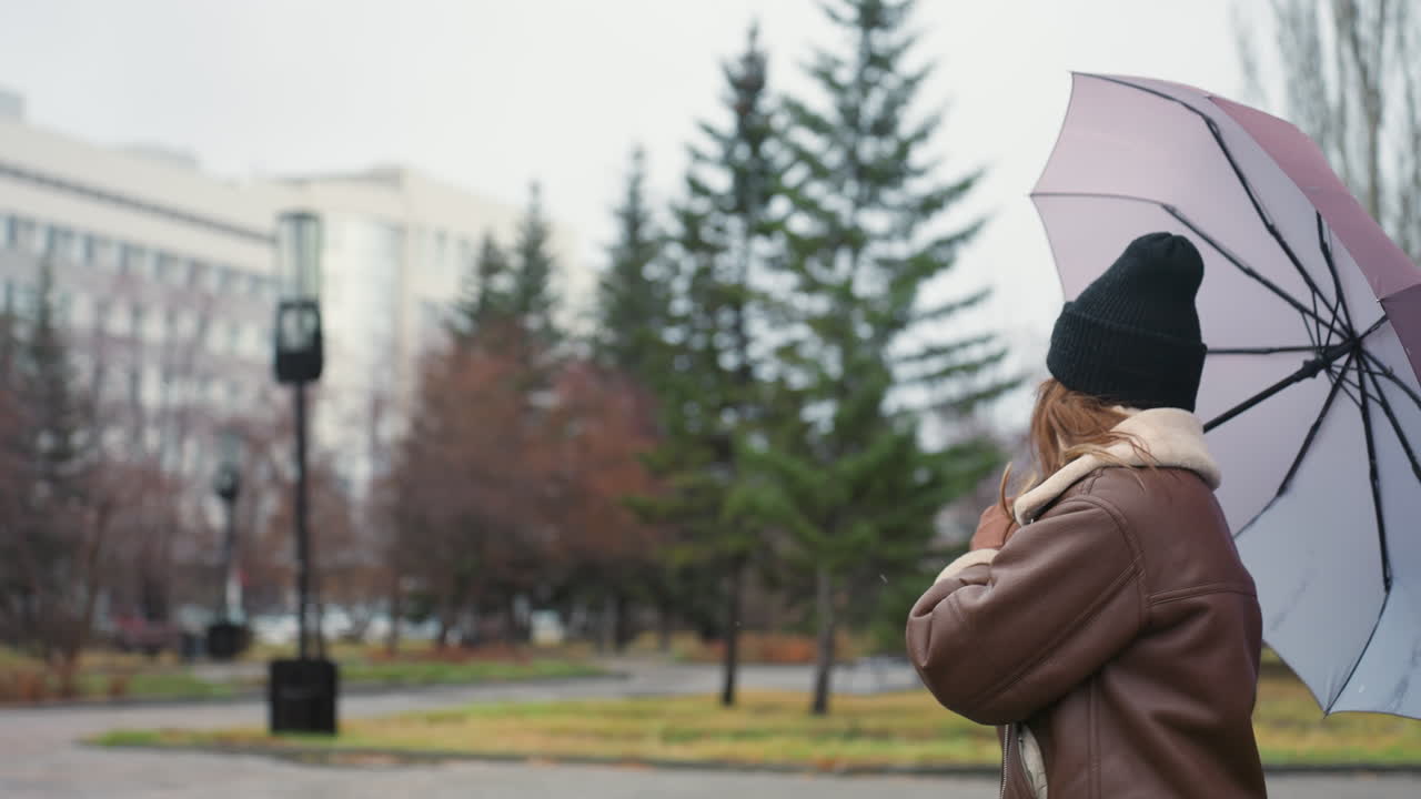Cheerful lady holding umbrella while walking in park on cold overcast day wearing black knit cap and brown shearling jacket, surrounded by pine trees and urban buildings with soft snowfall in background