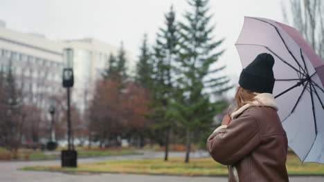 cheerful lady holding umbrella while walking in park on cold overcast day wearing black knit cap and brown shearling jacket, surrounded by pine trees and urban buildings with soft snowfall in background