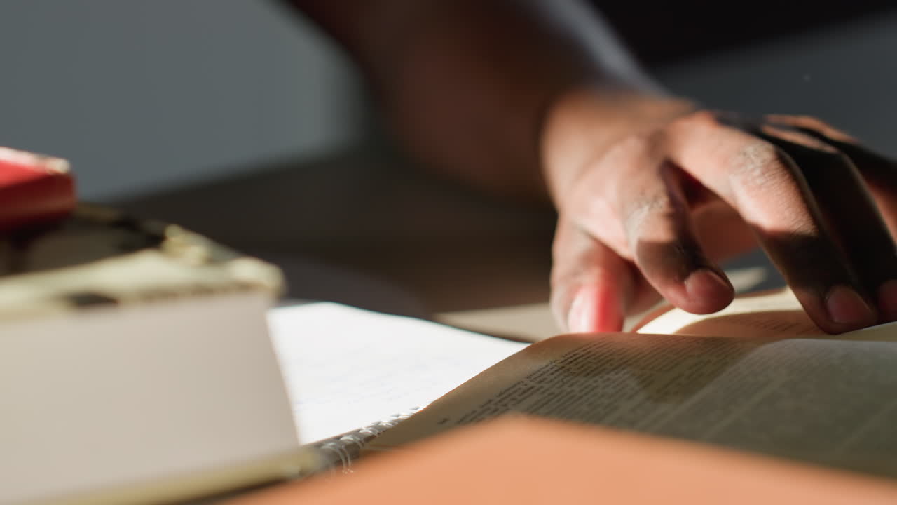 Close up of person opening page of book with fingers under warm light, showing textured paper, soft shadows, and focused moment of reading or studying in quiet indoor setting