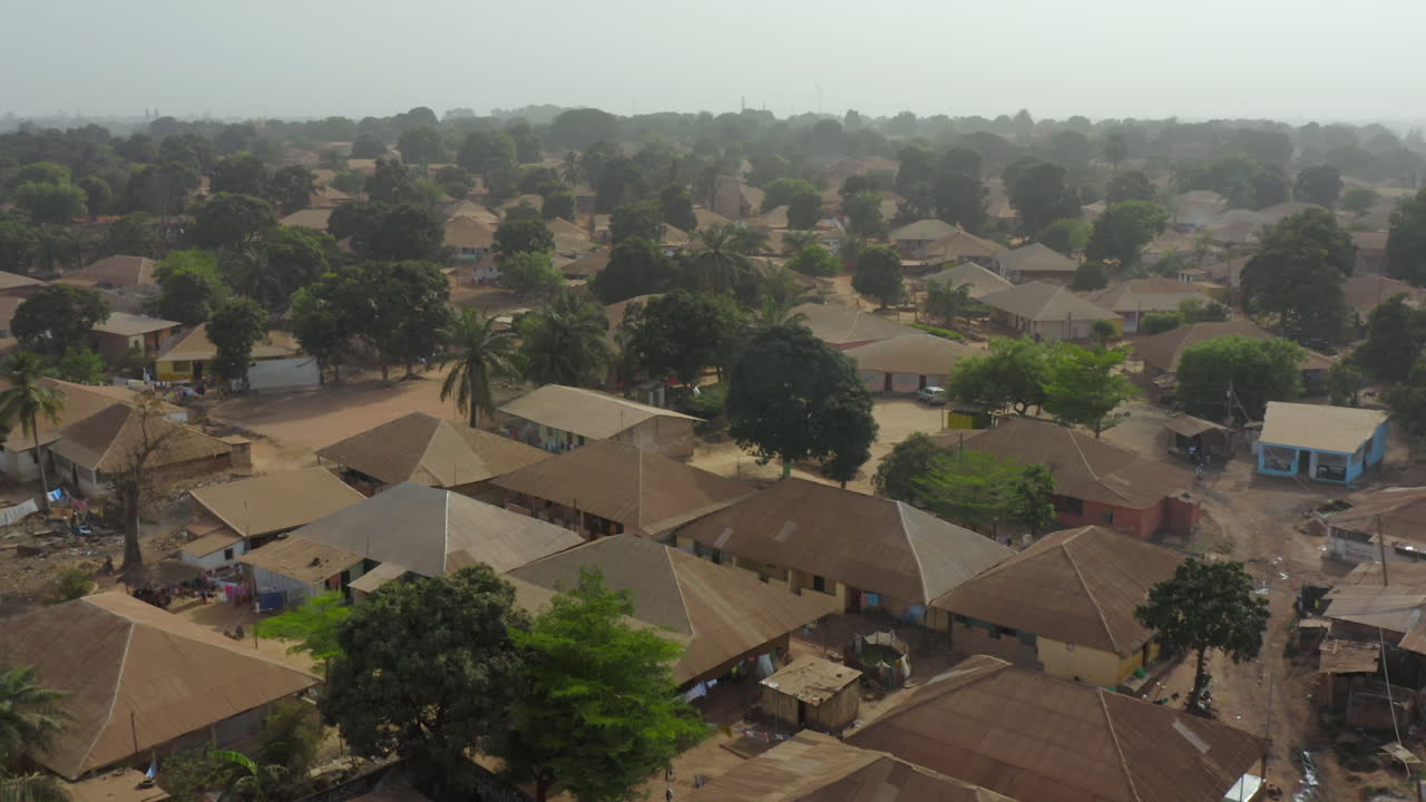 Drone shot focusing on the dense cluster of low-rise residential houses with red roofs, surrounded by trees and unpaved roads in a community in Guinea-Bissau, West Africa