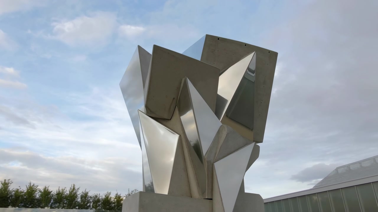 Vista em ângulo baixo de uma grande escultura contemporânea feita de blocos de concreto empilhados e facetas de metal espelhado polido, ambientada ao ar livre sob luz diurna suave e nublada, com forte contraste geométrico.