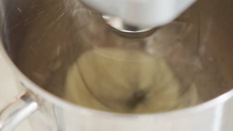 close up of electric mixer in motion as flour is poured into stainless steel bowl during baking preparation showing dynamic spinning blade and smooth metallic texture in bright kitchen environment
