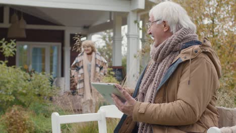 an older couple outside their home, the man uses a table while making a video call and invites his wife to join