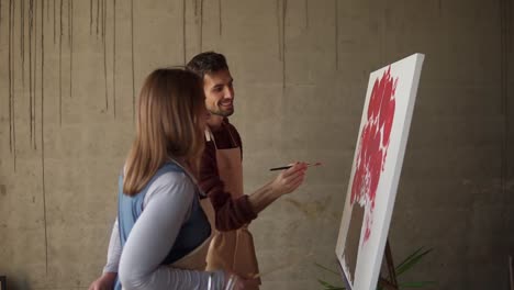 male student studying to paint with oil paints. dark haired man in beige apron drawing pink flowers using paintbrush under detailed explanation by female mentor. workshop in art studio