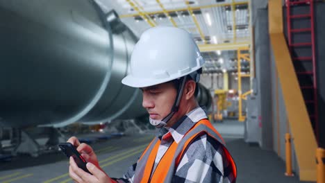 vista lateral de perto de um engenheiro masculino asiático com capacete de segurança usando um smartphone enquanto está de pé em uma fábrica de fabricação de tubos