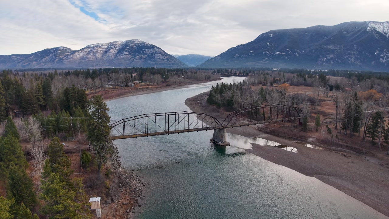 Wide aerial shot of a calm river meandering between forested banks, passing under a steel truss bridge with mountains in the background under a cloudy sky.