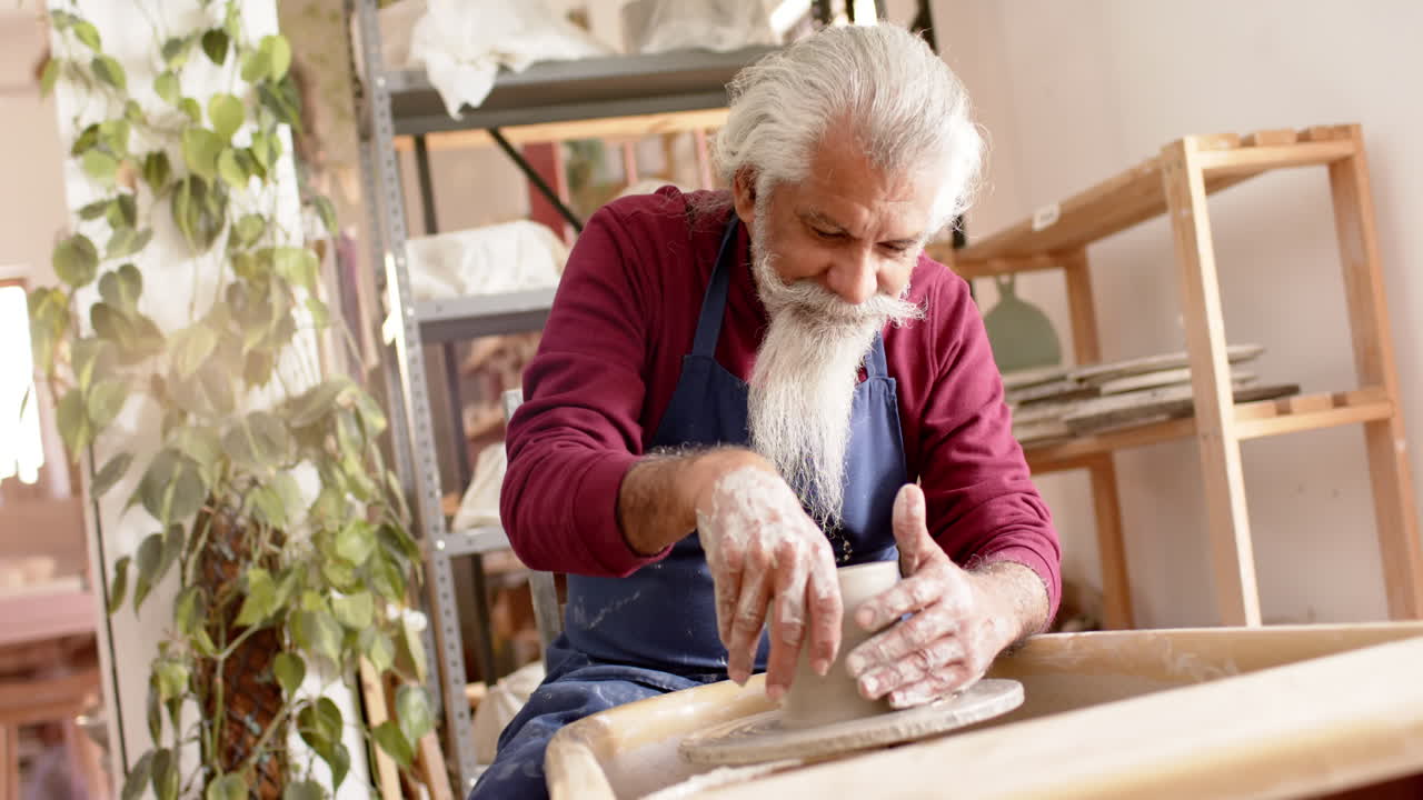 Anziano vasaio birazziale concentrato con barba lunga che usa il tornio in un laboratorio di ceramica, al rallentatore.