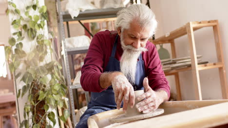 focused senior biracial potter with long beard using potter's wheel in pottery studio, slow motion