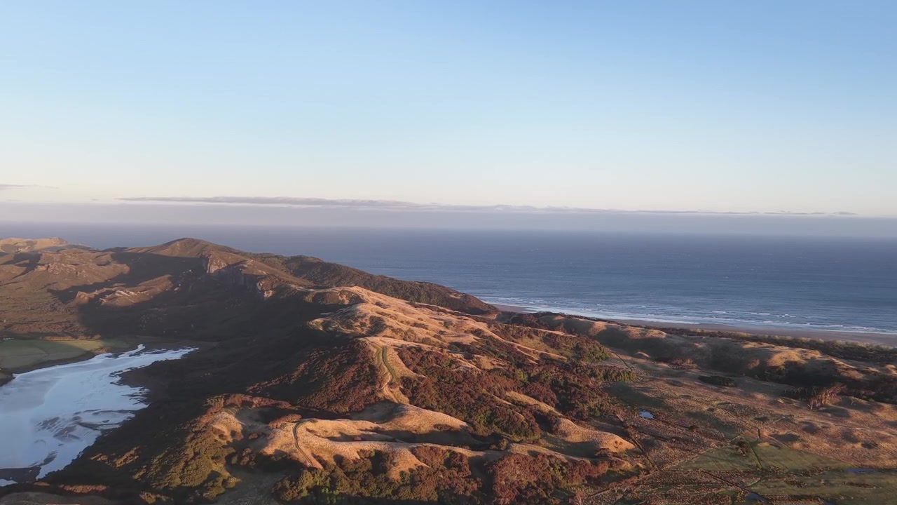 Filmatisk luftfoto av kystlinje og bølgende åser ved solnedgang, som viser dybde og naturlig landskap til bruk i reise og historiefortelling.