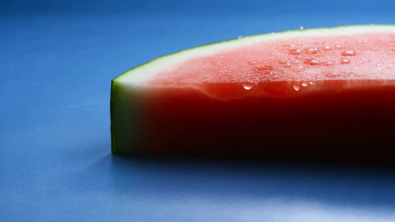 Un primo piano di una fetta di anguria fresca con buccia verde, polpa rossa e goccioline visibili, appoggiata su uno sfondo blu liscio in luce soffusa.