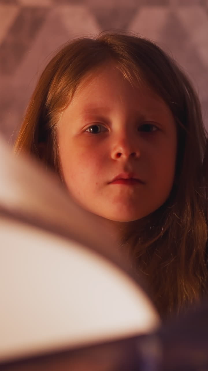 Attentive little girl looks at pages of fairy tale book turned by mother preparing to sleep at gentle light of night lamp in partially illuminated bedroom slow motion