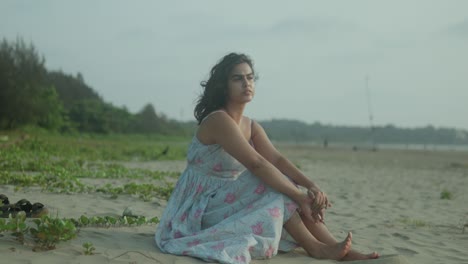 Woman-in-a-floral-dress-sitting-on-a-sandy-beach,-gazing-into-the-distance-with-greenery-and-trees-in-the-background