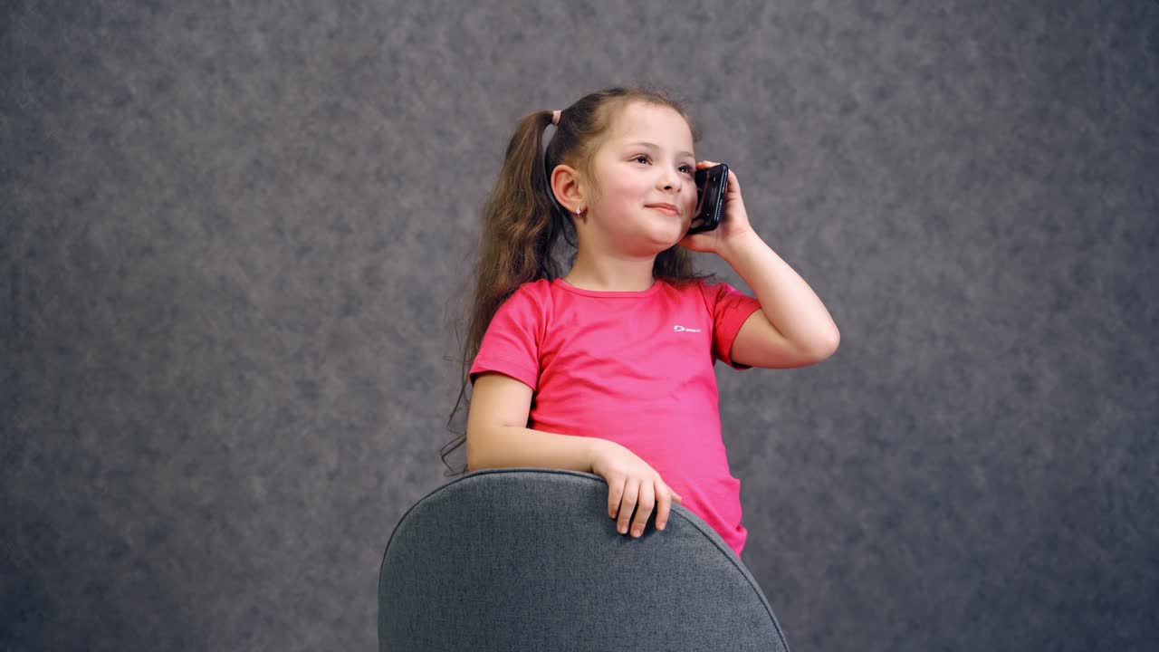 Positive little girl talking on the phone. Cheerful child in pink t-shirt on the chair answering the mobile phone. Beautiful little girl with a phone isolated on blue.