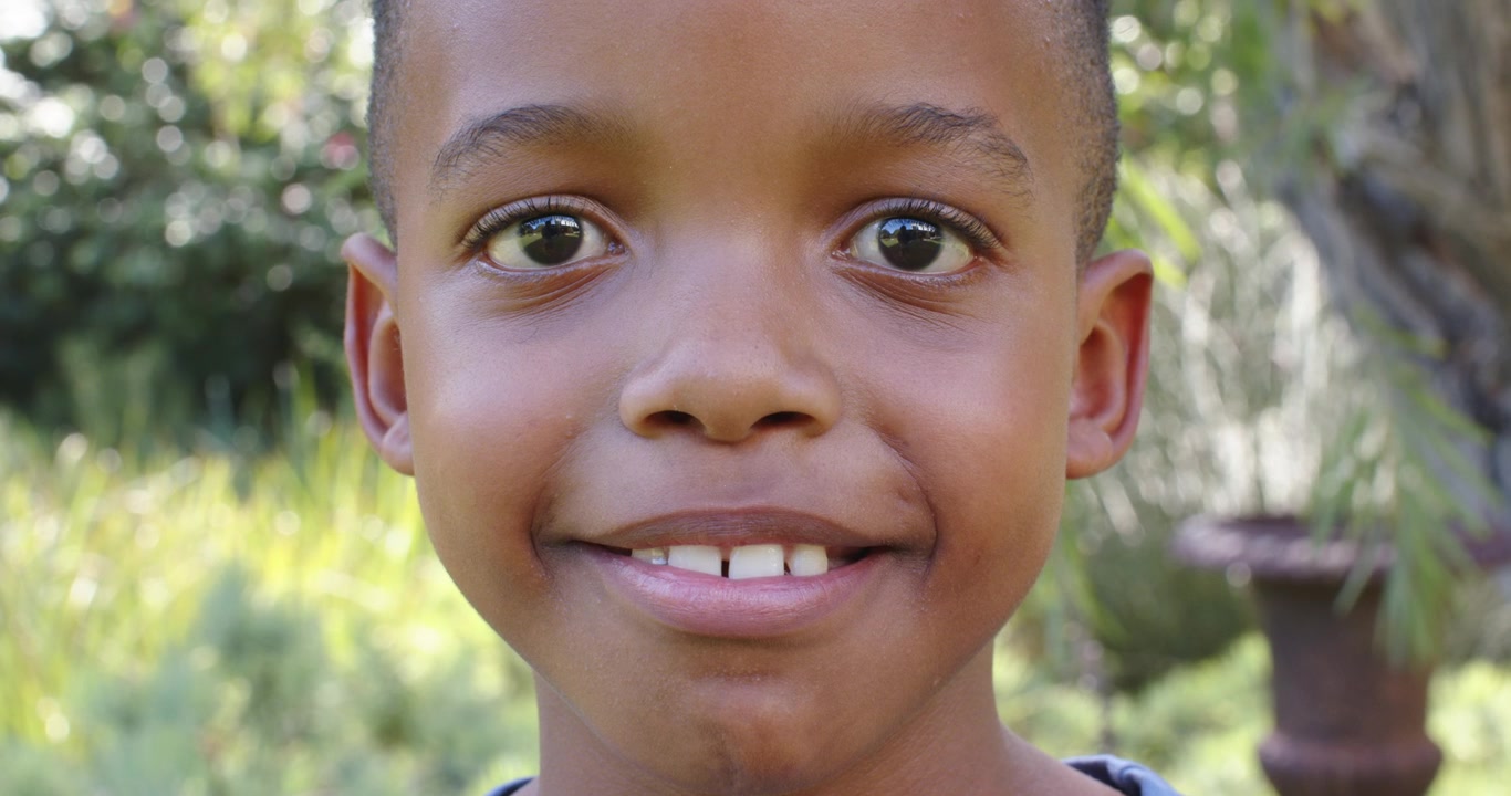 Bambino africano che risponde alla telecamera, con il viso rilassato, sorridendo in giardino vicino a un tronco d'albero e un'urna.