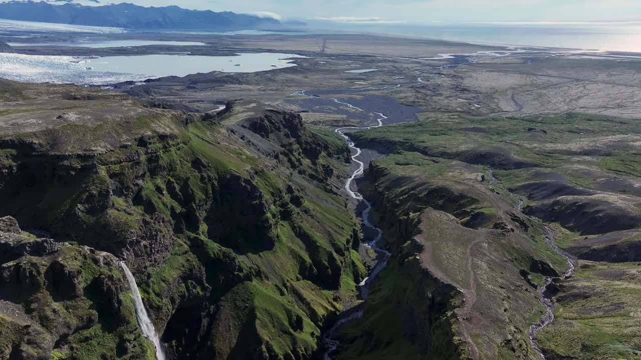 Aerial establishing shot of Mulagljufur canyon on Iceland island. Flowing river into valley. Glacier mountains in background. Falling waterfall down rocky Canyon. Wide shot