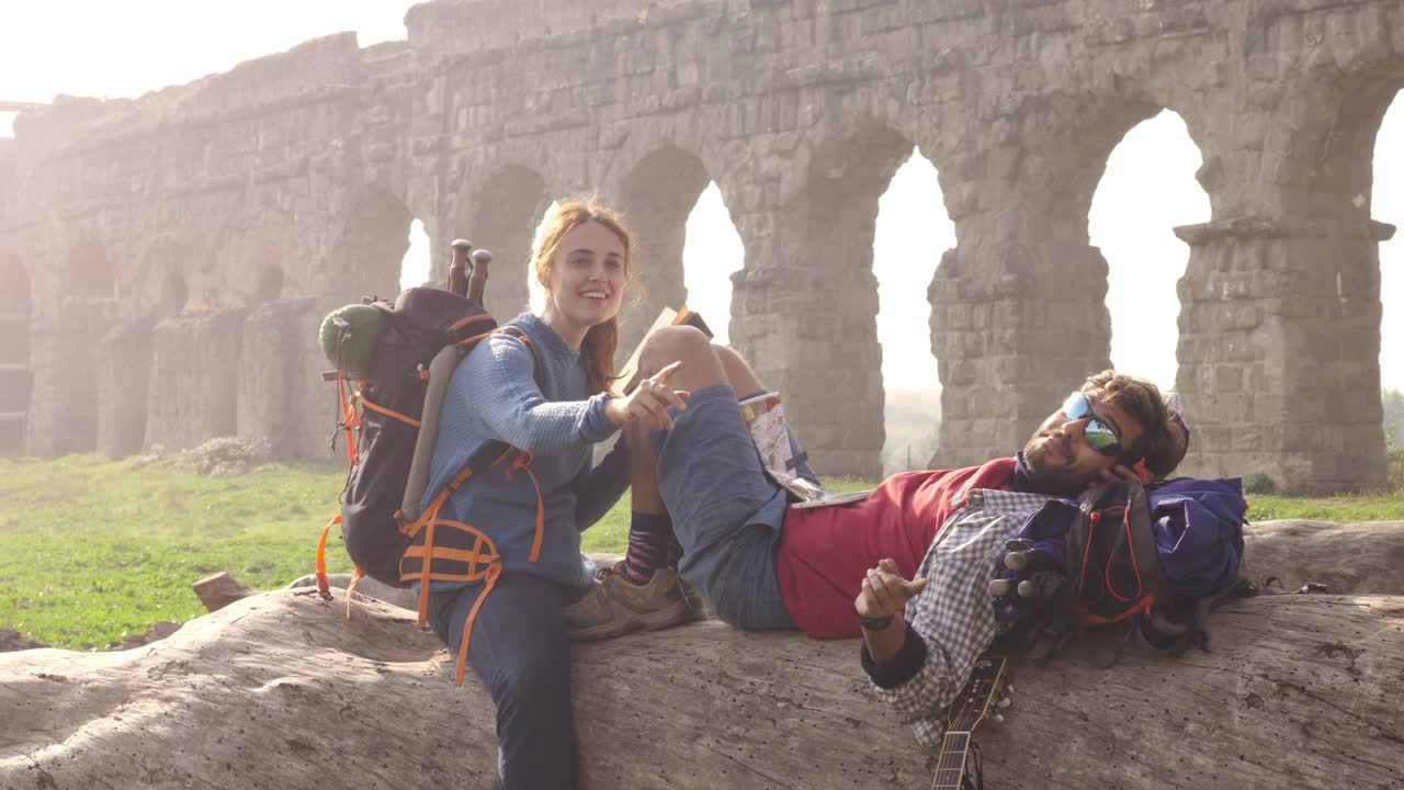 una pareja feliz de mochileros turistas sentados acostados en un tronco de madera leyendo un libro usando un teléfono inteligente frente a las antiguas ruinas del acueducto romano en el parque parco degli aquedotti en roma al amanecer romántico con saco de dormir en cámara lenta