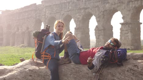 happy young couple backpackers tourists sitting lying on a log trunk reading book using smartphone in front of ancient roman aqueduct ruins in parco degli acquedotti park in rome at sunrise romantic with sleeping bag slow motion