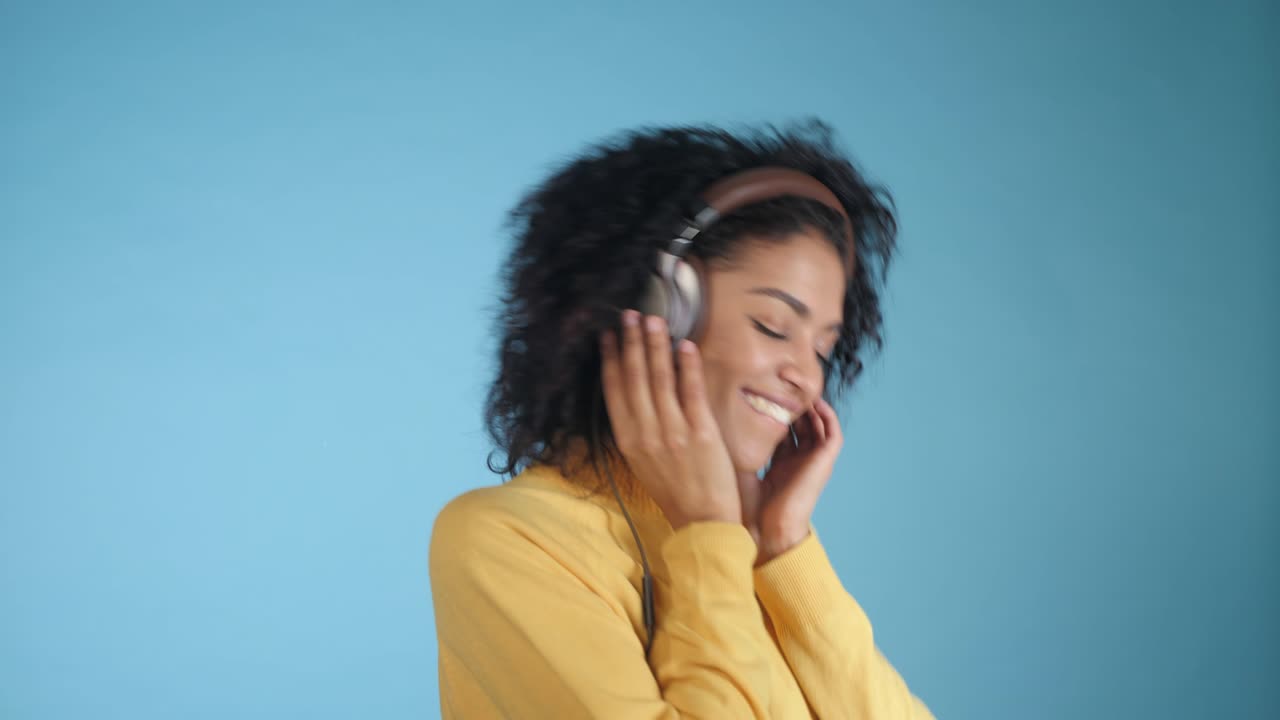 Mannequin souriant 4K avec un casque audio posant et dansant sur fond bleu en studio. Belle jeune mannequin femme métisse africaine appréciant la musique