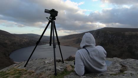 enthusiastic photographer making time lapse at lake guinness in ireland, sitting on rocks with hoodie next to hes camera on tripod