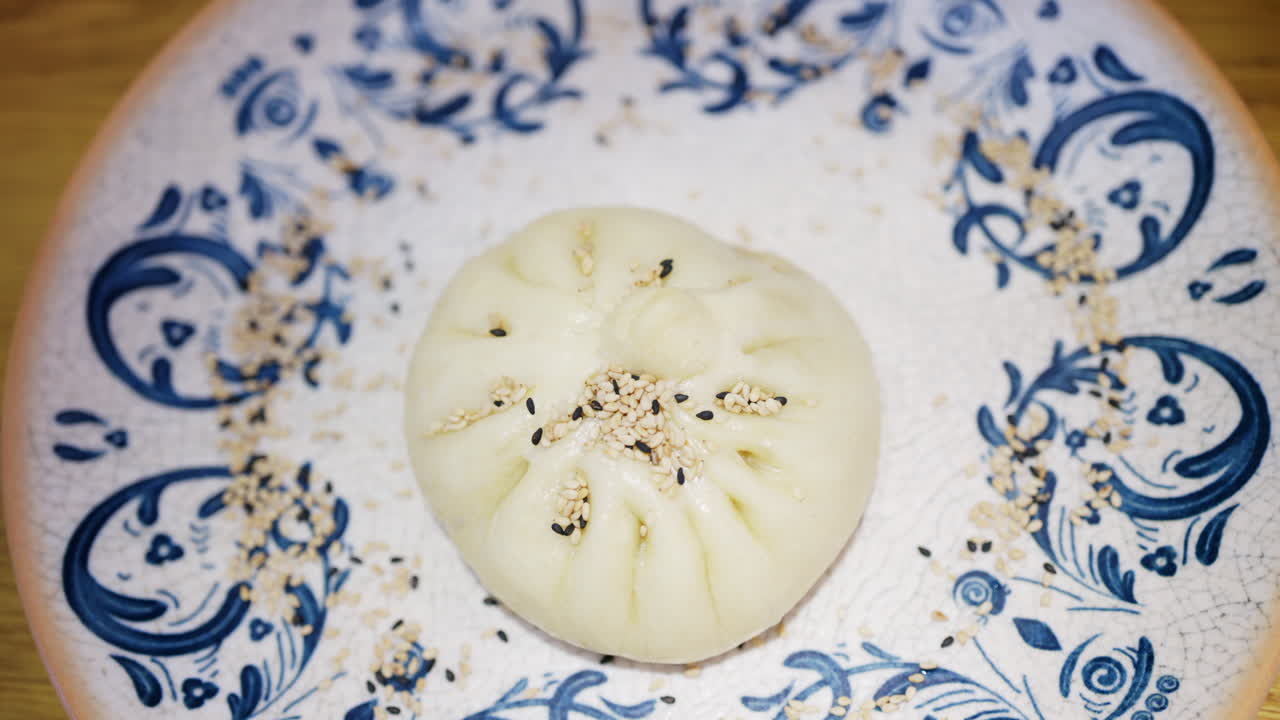 Primo piano di un panino al vapore guarnito con semi di sesamo su un piatto di ceramica decorativo.