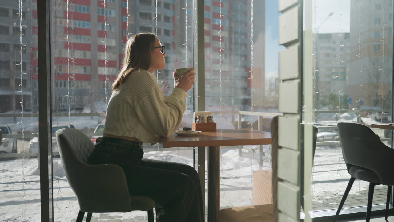 Young professional woman sitting inside cafe enjoying tea by window on sunny winter day, cars parked and passing outside, urban buildings and street scene visible through glass