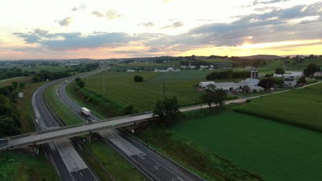 cars and trucks, on highway under dramatic sunrise, sunset among green rural american farmland during summer, rising aerial of traffic