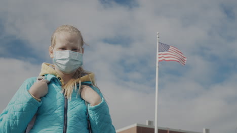 a child in a protective mask stands against the background of a school and a us flag.