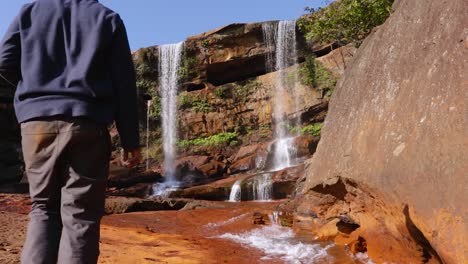 Joven-Caminando-Hacia-La-Prístina-Cascada-Natural-Que-Cae-Desde-La-Cima-De-La-Montaña-En-El-Día-Desde-Un-Video-De-ángulo-Bajo-Tomado-En-Phe-Phe-Fall-Meghalaya-India