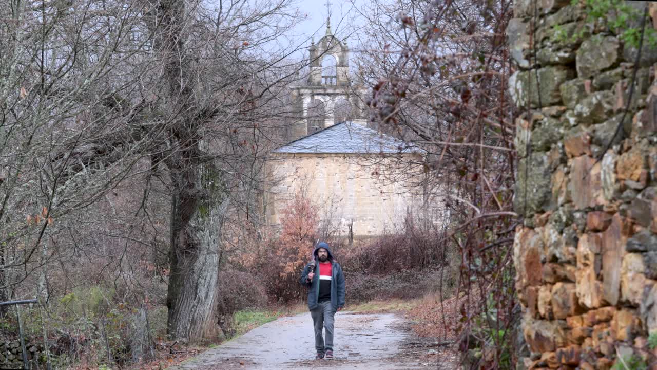 un joven sale de una iglesia rural en medio de la naturaleza después de un día lluvioso con un paraguas cerrado en la mano