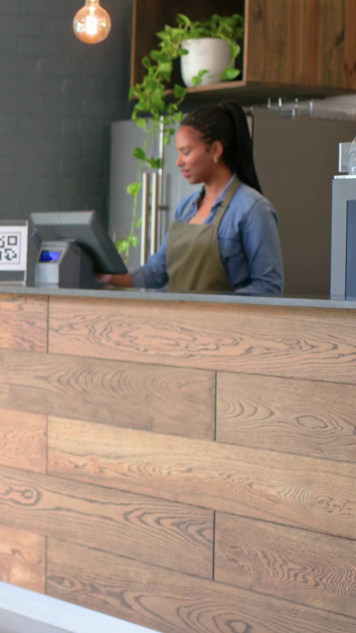 Video vertical: Mujer saludando con top de mezclilla y delantal verde oliva, usando un TPV para procesar una venta en el mostrador con un código QR.