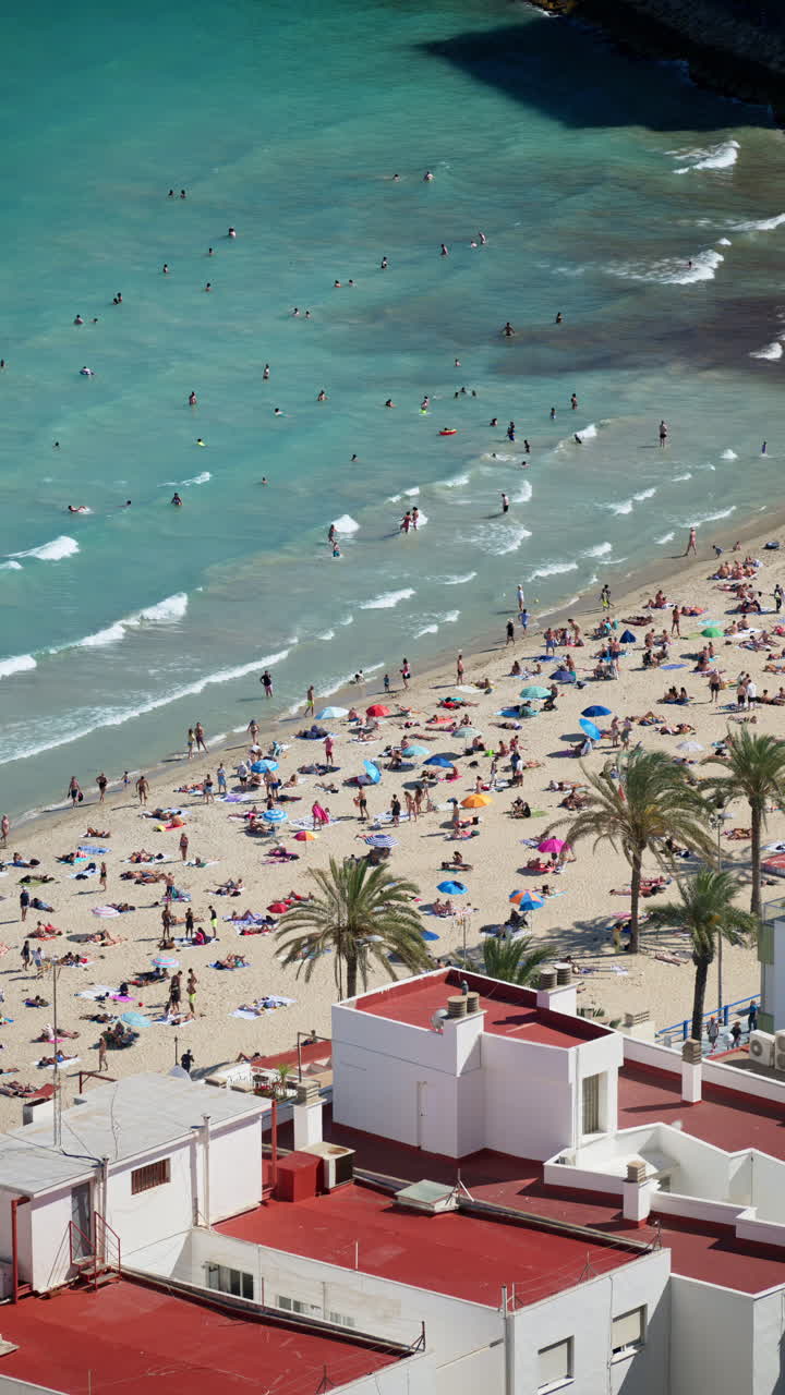 Aerial view of turquoise water, a sandy beach and seafront apartment blocks with swimmers and umbrellas visible below. Vertical