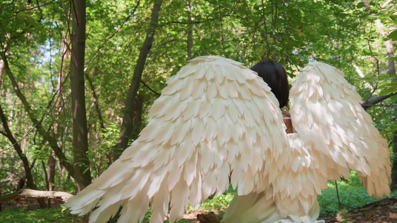 Rear view fairy guardian creature with majestic white wings sitting gracefully on fallen tree amid lush green forest under warm sunlight creating magical fantasy