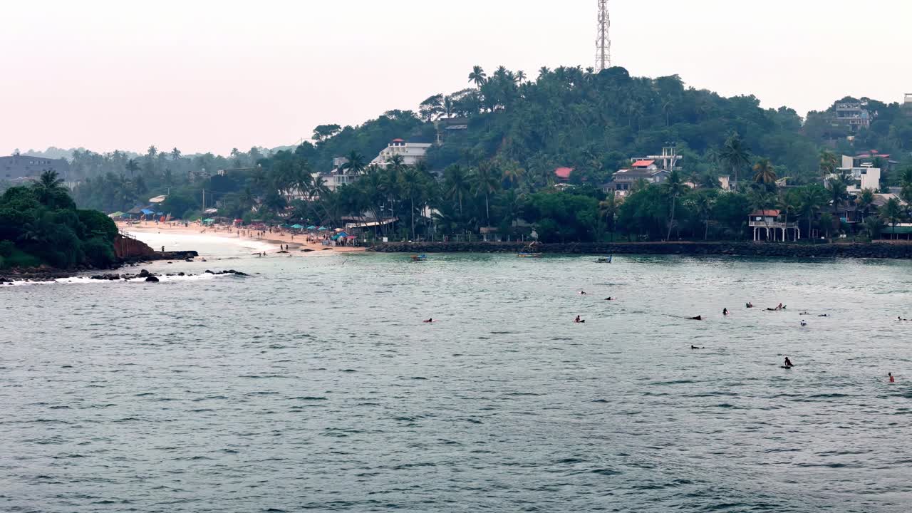 Aerial Elevated wide shot of Sri Lankan bay on a hazy day Swimmers enjoy calm grey waters near beach lush hill crowned with buildings tower Standard lens view