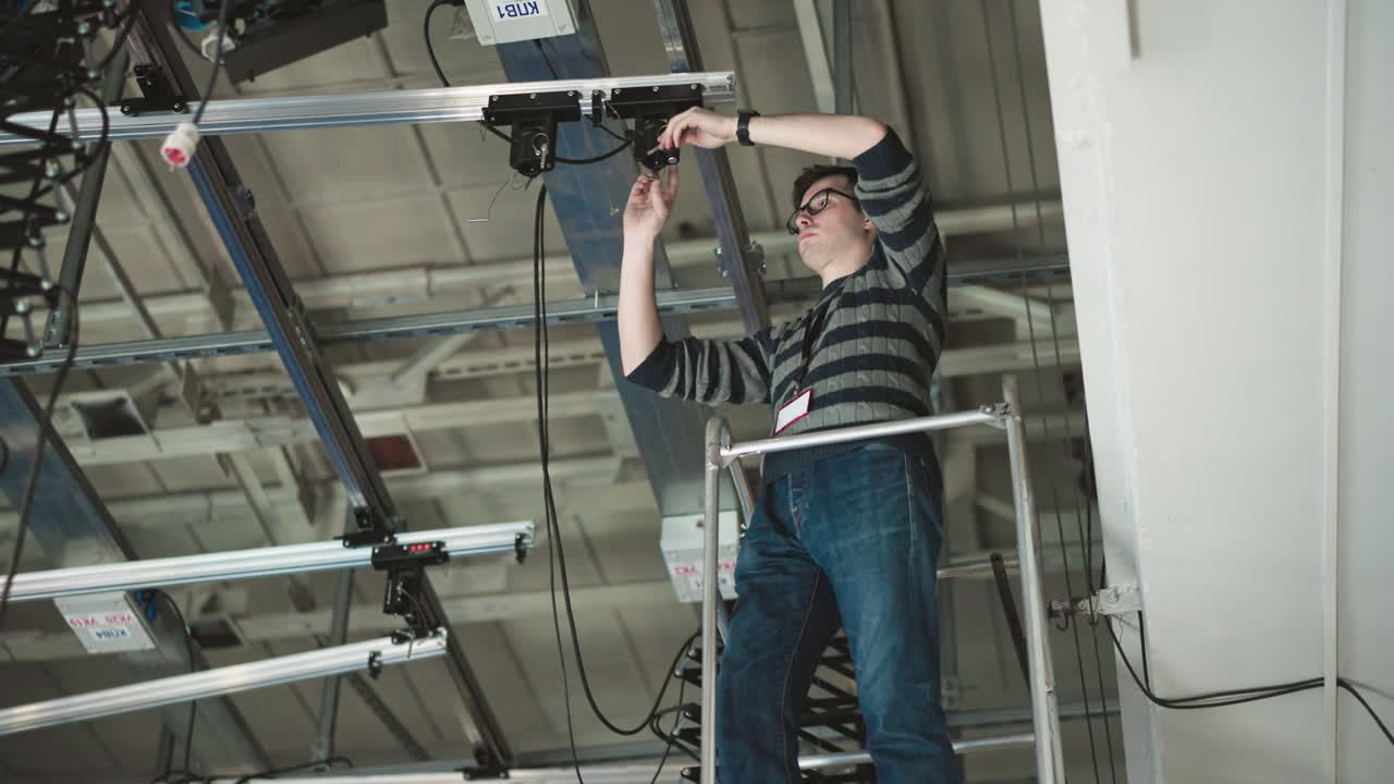 Person working on elevated platform, adjusting camera or lighting equipment mounted on ceiling grid, wearing sweater and glasses, visible cables, industrial setting, maintenance or installation task